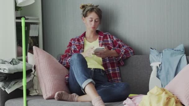 Güzel genç bayan temizlik yaptıktan sonra kanepede dinleniyor ve telefonu kullanıyor. — Stok video