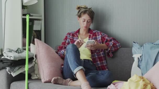 Όμορφη νεαρή γυναίκα ξεκουράζεται στον καναπέ μετά τον καθαρισμό και τη χρήση του τηλεφώνου — Αρχείο Βίντεο