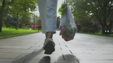Öndeki kadın elinde sebzelerle pamuk örgüsü alışveriş çantası tutuyor ve yaz parkında yürüyor.