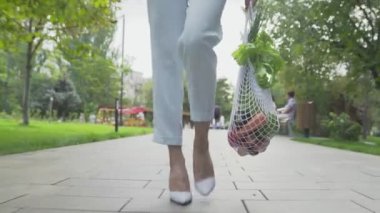 Mavi pantolonlu ve beyaz ayakkabılı genç bir kadın, elinde sebzelerle pamuk örgüsü alışveriş torbası tutuyor ve yaz parkında yürüyor. Sıfır atık.