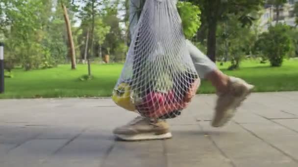 Oigenkännlig kvinna håller bomull mesh shoppingväska med grönsaker och promenader i sommarparken — Stockvideo