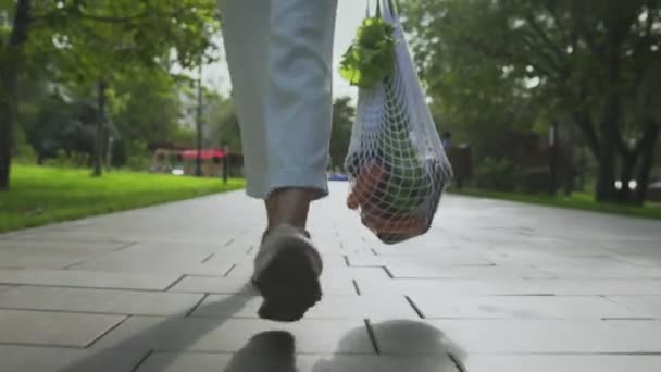 Framsidan av kvinnan håller bomull mesh shoppingväska med grönsaker och promenader i sommarparken — Stockvideo