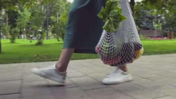 Sidovy över ung kvinna i grön kjol och vita sneakers håller bomull mesh shoppingväska med grönsaker och promenader i sommarparken. Handeln med nollavfall. — Stockvideo