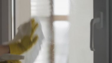 Sarı eldivenli ev kadını camları beyaz bez ve deterjanla yıkıyor..