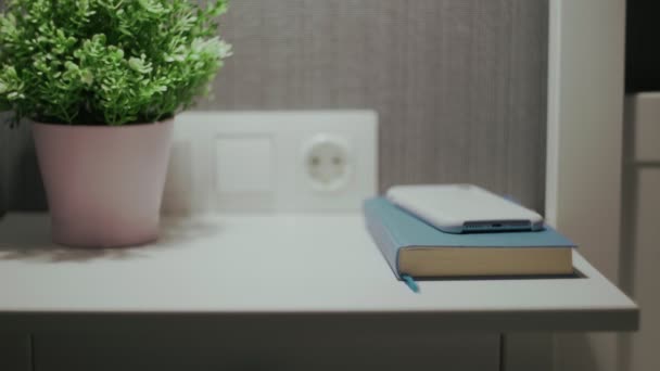 Female hand turn off an alarm clock on mobile phone on bedside table with indoor plant and book — Stock Video