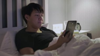 Tablet bilgisayarlı genç Asyalı adam yatakta yatıyor.