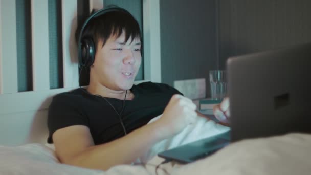 Joven asiático hombre en auriculares usando un portátil tarde en la noche en la cama — Vídeos de Stock