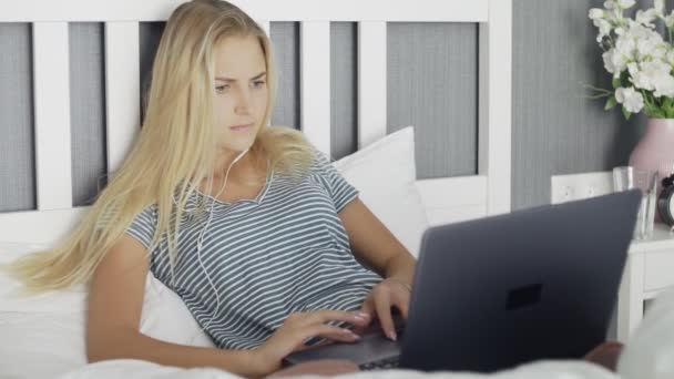 Kulaklıklı genç bir kadın internette dizüstü bilgisayar kullanarak yatağa uzanıyor.. — Stok video