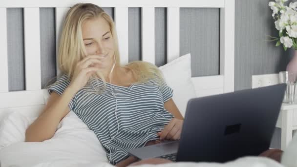 Mujer joven feliz con auriculares tumbados en la cama usando el ordenador portátil para ver un video divertido — Vídeos de Stock