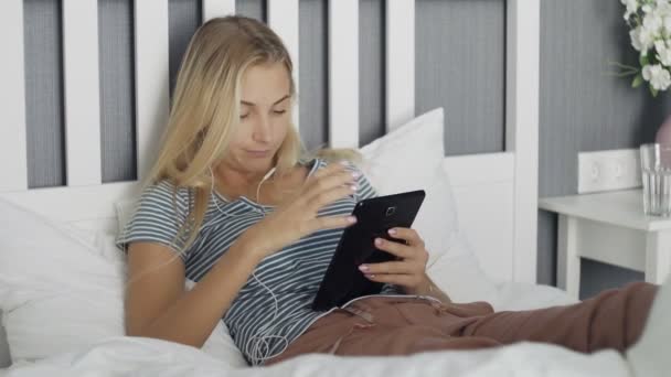 Jovem mulher entediada navegando nas mídias sociais em um tablet deitado em uma cama em casa — Vídeo de Stock