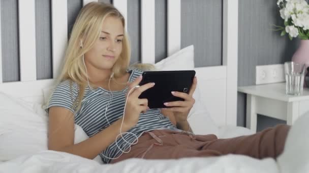 Mujer joven viendo los medios de comunicación en una tableta acostada en una cama en casa — Vídeos de Stock