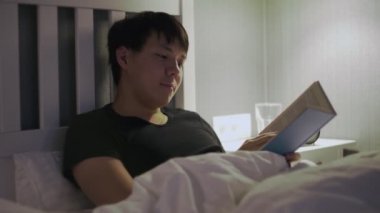 Genç Asyalı adam yatakta uzanıp kitap okuyor.