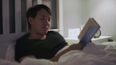 Genç Asyalı adam yatakta uzanıp kitap okuyor.