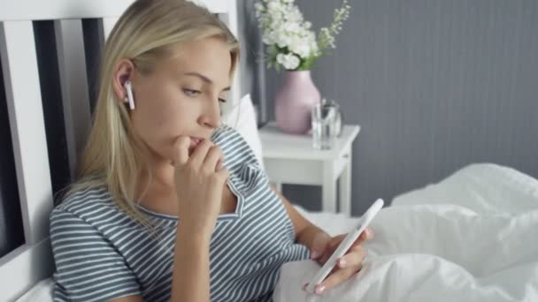 Mujer joven hermosa feliz con auriculares inalámbricos tumbados en la cama y el uso de teléfono inteligente — Vídeos de Stock