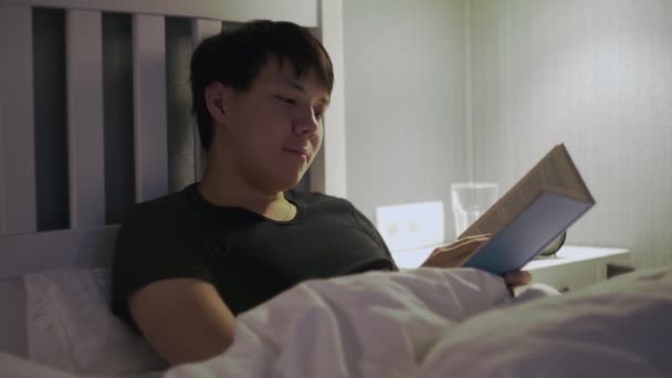 Jonge aziatische man liggend in bed en het lezen van een boek — Stockvideo