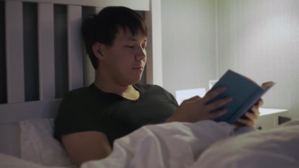 Молодой человек лежит в постели и читает книгу, а потом засыпает. — стоковое видео