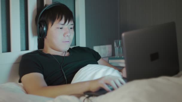 Νεαρός Ασιάτης με ακουστικά που χρησιμοποιεί φορητό υπολογιστή αργά τη νύχτα στο κρεβάτι — Αρχείο Βίντεο