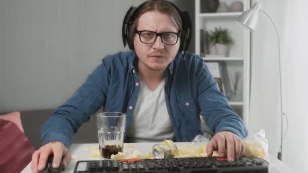Сконцентрированный молодой человек играет в видеоигры, ест чипсы и пьет колу — стоковое видео