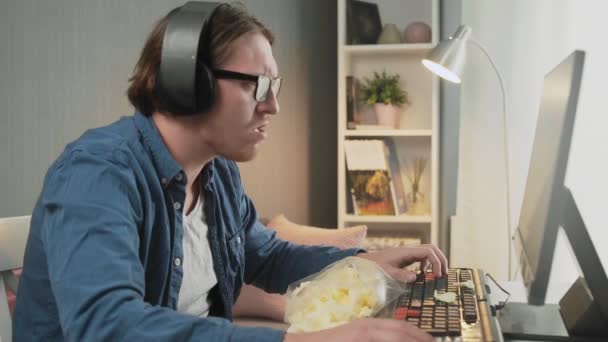 Vista laterale del giovane con le cuffie che giocano al videogioco e mangiano patatine a casa — Video Stock