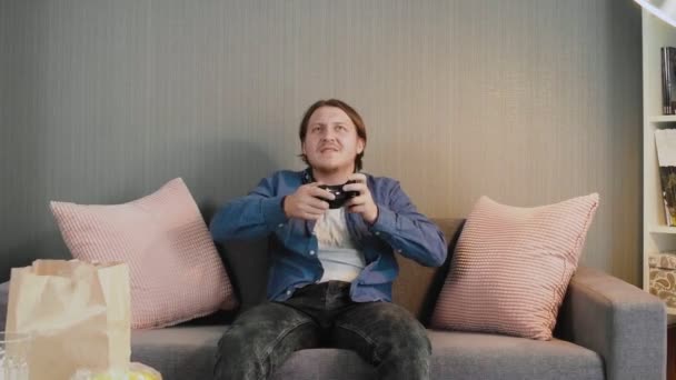 Молодой геймер, держащий джойстик и играющий в видеоигры, сидит дома один на диване — стоковое видео