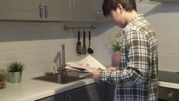 Schläfrig asiatisch mann mit zeitung taking ein cookie zuhause im die küche. — Stockvideo