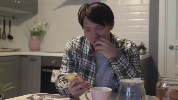 快乐的亚洲人在智能手机上读着有趣的东西，在家里的厨房里笑着 — 图库视频影像