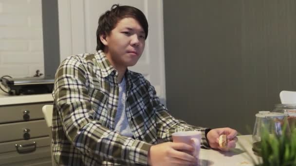 Sleepy asian man drinking coffee and eating cookie at the table. — Stock Video