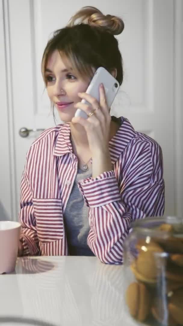 Vídeo vertical de una joven sonriente tomando un café y hablando por teléfono en la cocina — Vídeos de Stock