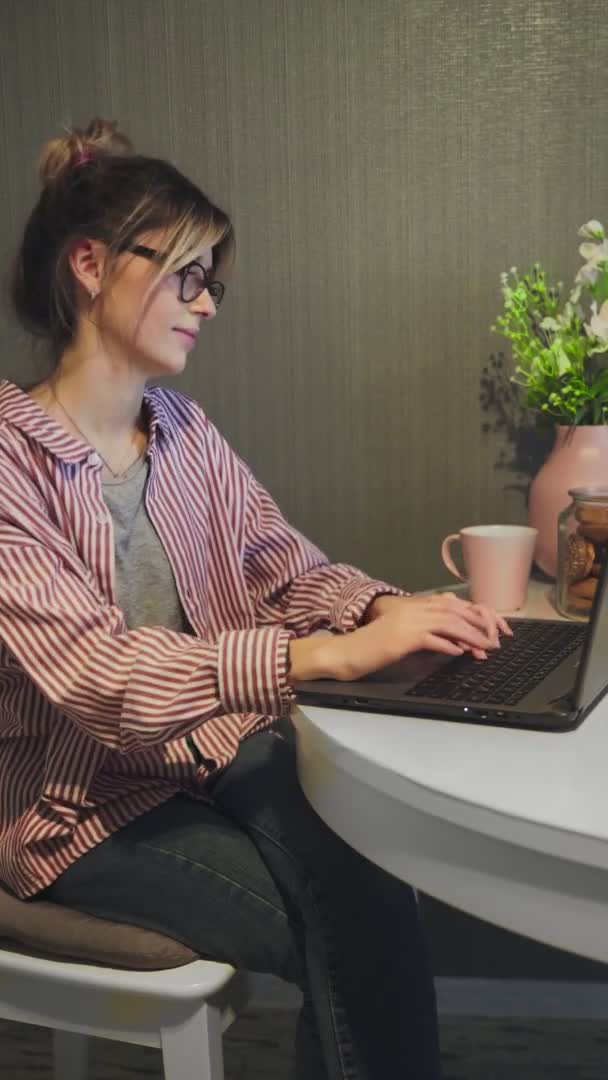 Вертикальное видео молодой женщины сидит за столом на кухне и типы на ноутбуке — стоковое видео