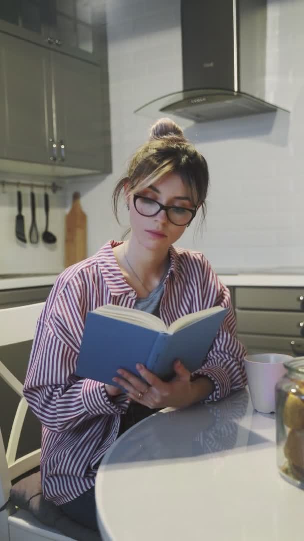 Κάθετη βίντεο από νεαρή όμορφη μελαχρινή γυναίκα πίνει καφέ και διαβάζει βιβλίο στο σπίτι στην κουζίνα — Αρχείο Βίντεο