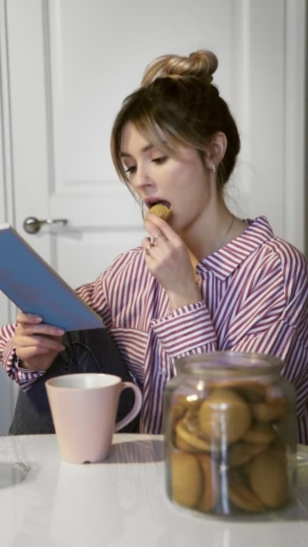 Вертикальное видео, где молодая красивая брюнетка пьет кофе, ест печенье и читает книгу дома на кухне — стоковое видео
