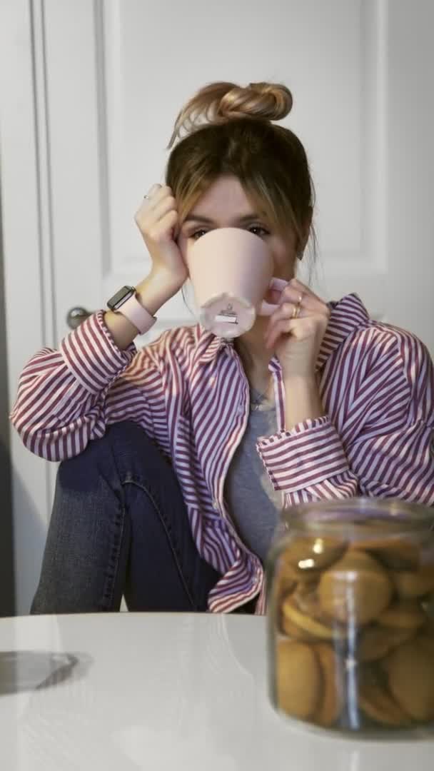Vertikal video av ung vacker brunett kvinna dricker kaffe och äter kaka hemma i köket — Stockvideo