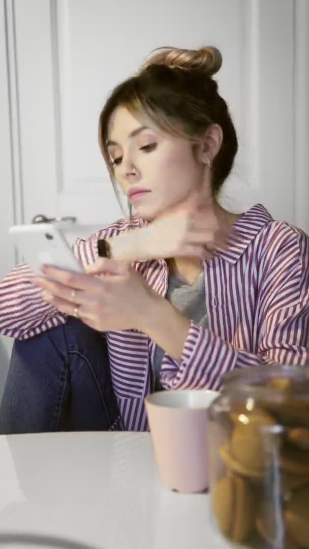 El vídeo vertical de la muchacha joven por la mañana en la cocina usando el teléfono — Vídeos de Stock