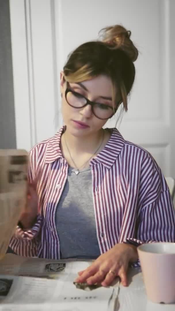 Vertical video of young beautiful brunette woman with glasses drinks coffee, eats cookie and reads newspaper at home in kitchen — Stock Video