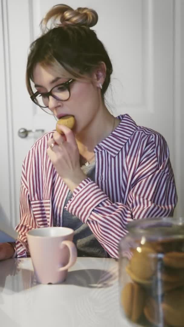 Vertical video of young beautiful woman with glasses drinks coffee, eats cookie and reads book at home in kitchen — Stock Video