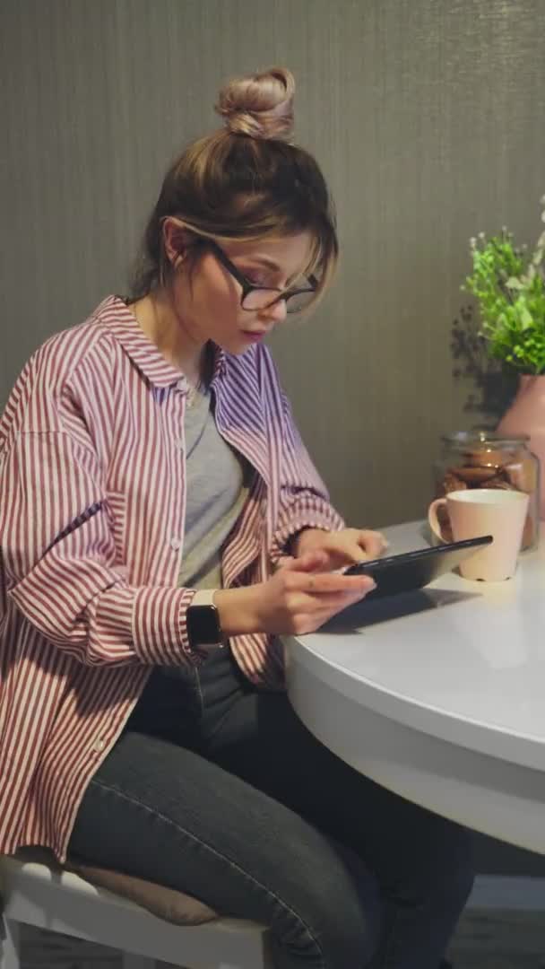Вертикальне відео привабливої жінки в окулярах, що друкуються на цифровому планшеті на кухні — стокове відео
