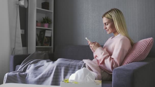 Mutlu ve rahat genç bir kadın cep telefonu ekranına bakıp gülüyor alışveriş için mobil uygulamalar kullanıyor oyun oynuyor sohbet ediyor sosyal medyada sohbet ediyor evde kanepede oturuyor. — Stok video