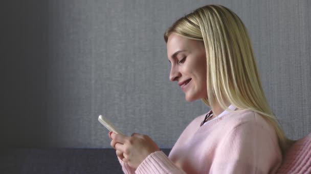 Красивая молодая женщина в розовом свитере держит в руках смартфон и печатает на его экране, разговаривая в гостиной дома — стоковое видео