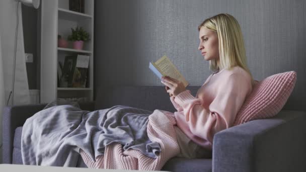 Mujer atractiva blanca leyendo un libro en el sofá gris en la acogedora sala de estar — Vídeos de Stock