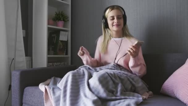 Ung kvinna i hörlurar lyssnar glad musik och dansar på soffan — Stockvideo