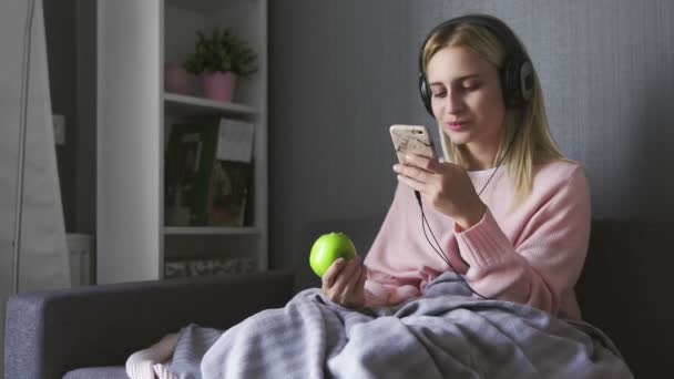 Молодая женщина в наушниках слушает счастливую музыку и ест яблоко — стоковое видео