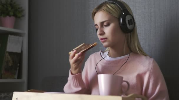 Mujer joven se sienta en el sofá come tostadas con pasta de chocolate y escuchar la música en el teléfono inteligente — Vídeo de stock