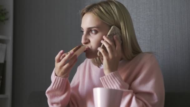 Junge Frau sitzt auf Couch isst Toast mit Schokopaste und telefoniert — Stockvideo