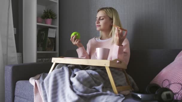 Ung kvinna sitter på soffan och gör ett val vad man ska äta rostat bröd med choklad eller äpple — Stockvideo