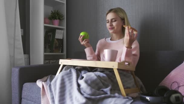 Mujer joven se sienta en el sofá y hace una elección qué comer tostadas con chocolate o manzana — Vídeos de Stock