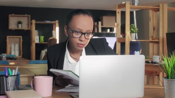 Jeune femme afro-américaine tapant sur un ordinateur portable au bureau puis écrivant dans un cahier de travail — Video