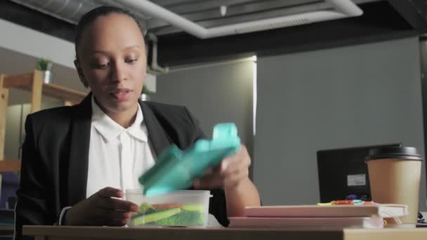 Vista lateral de una joven afroamericana almorzando en la oficina. Mujer come verduras de contenedor — Vídeos de Stock