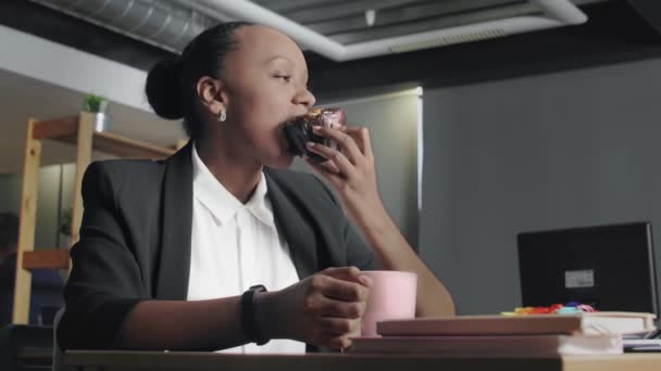 Vista de una mujer afroamericana almorzando en la oficina. Mujer come panecillo y bebe un café — Vídeos de Stock