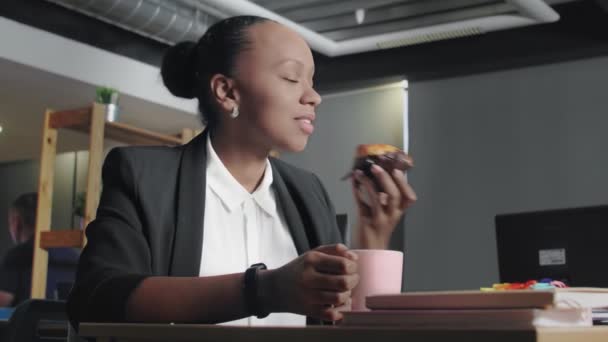 Vista de una mujer afroamericana almorzando en la oficina. Mujer come panecillo y bebe un café — Vídeos de Stock