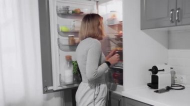 Kadın mutfaktaki buzdolabının kapısını açıyor ve patates kızartması yiyor.